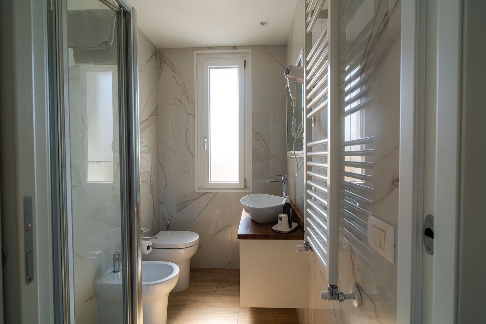 Ein Badezimmer mit Duschabtrennung aus Glas, Toilette, Waschbecken mit Marmorplatte und einem Fenster, das für natürliches Licht sorgt.