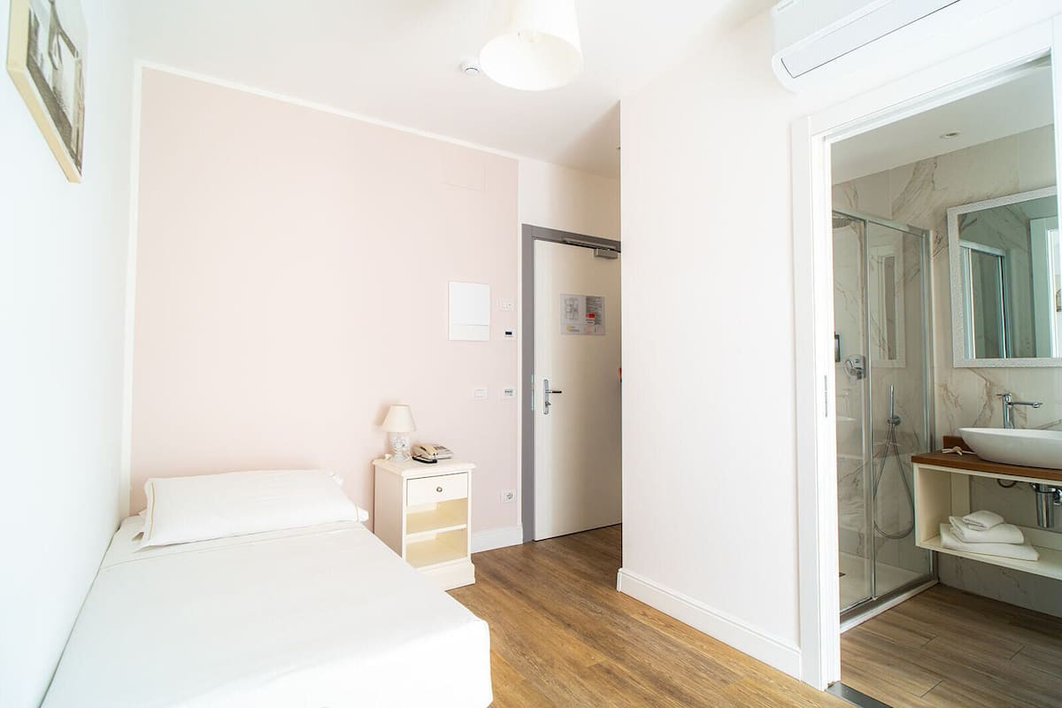 A bedroom with a bed, bedside table, lamp, and a door leading to a bathroom with a shower and sink.