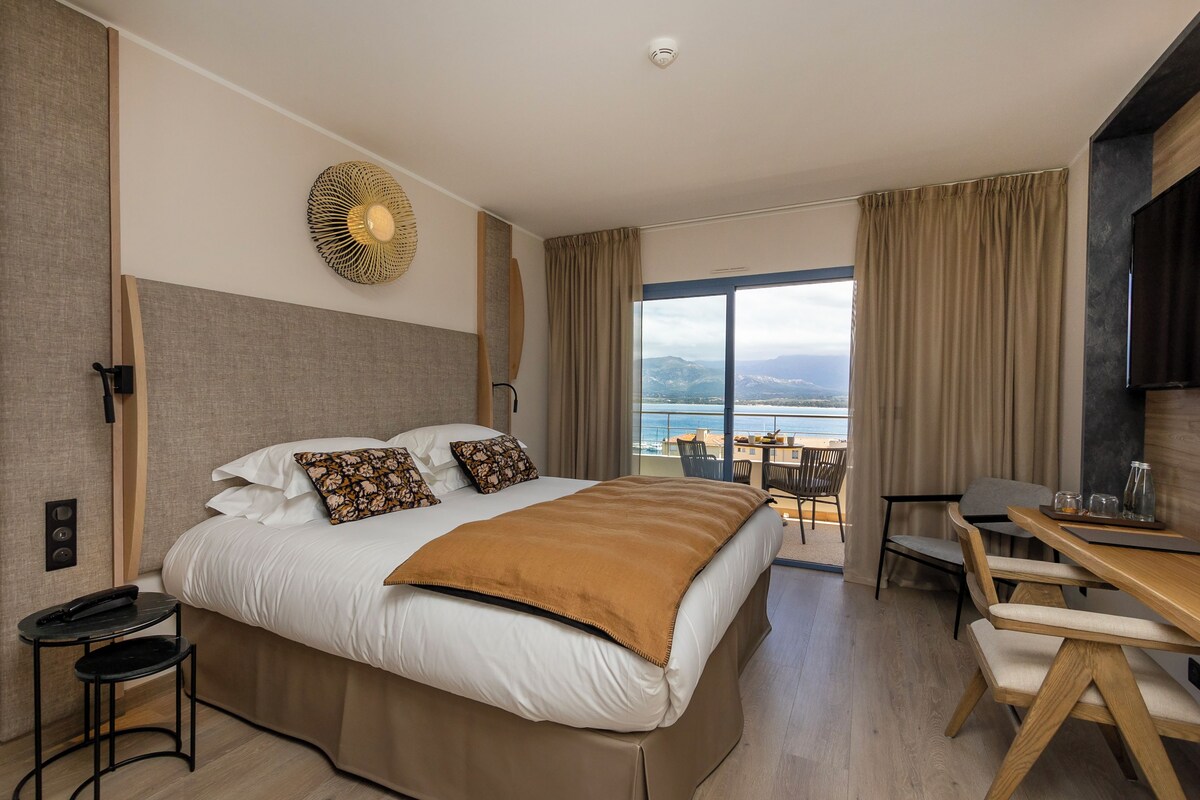 A modern hotel room with a large bed, a desk, and a view of the sea.