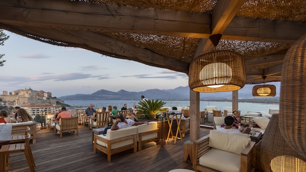 Une terrasse en bois avec des sièges extérieurs, offrant une vue sur une étendue d’eau et sur des montagnes lointaines.