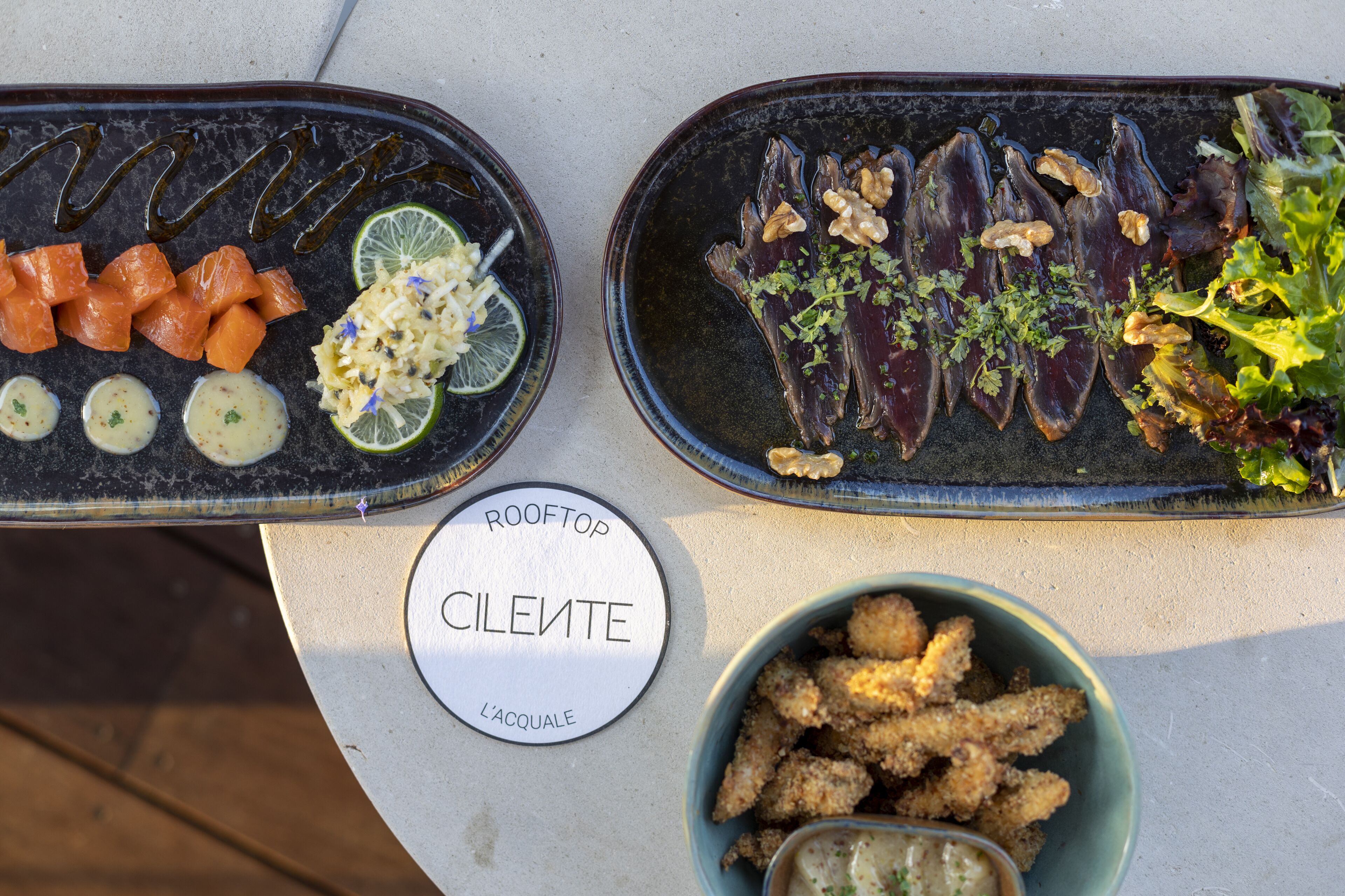 A table with two platters of food, a bowl of fried items, and a sign with the name 'ROOFTOP CLEMENTE'.