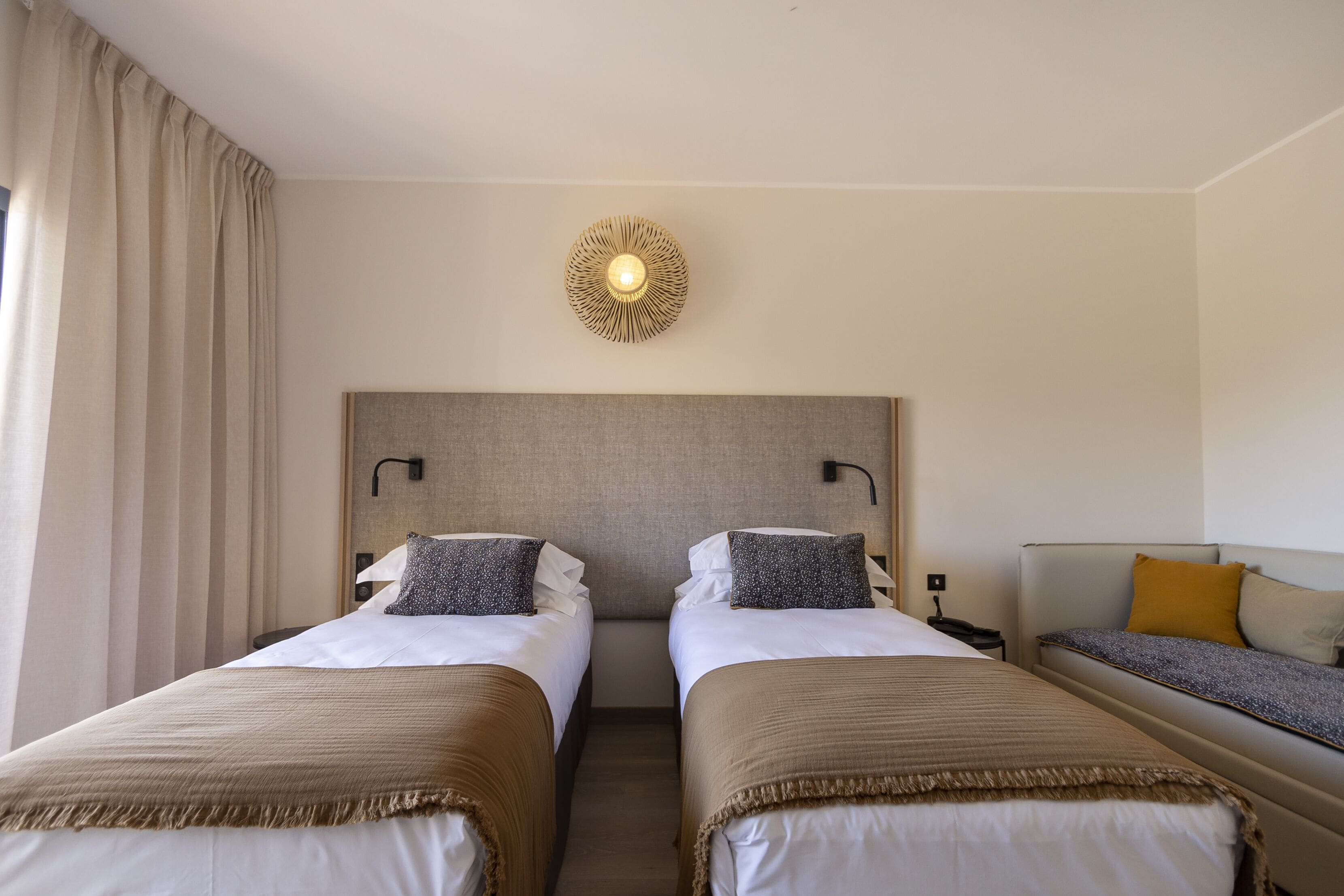 A hotel room with two single beds, a sofa, a wall-mounted light fixture, and a curtain on the window.