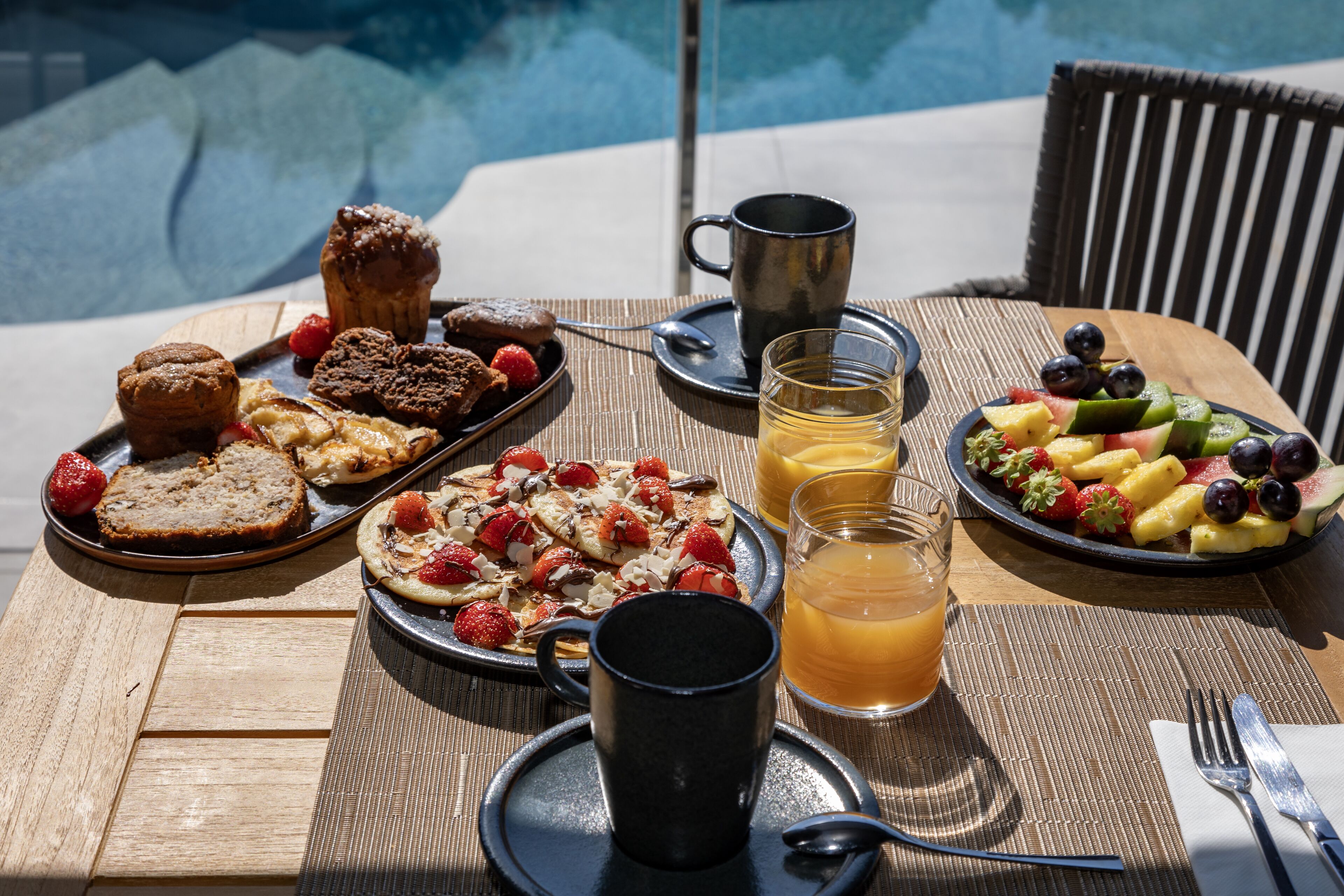 Desayuno con pastelería, frutas y bebidas sobre una mesa junto a una piscina.