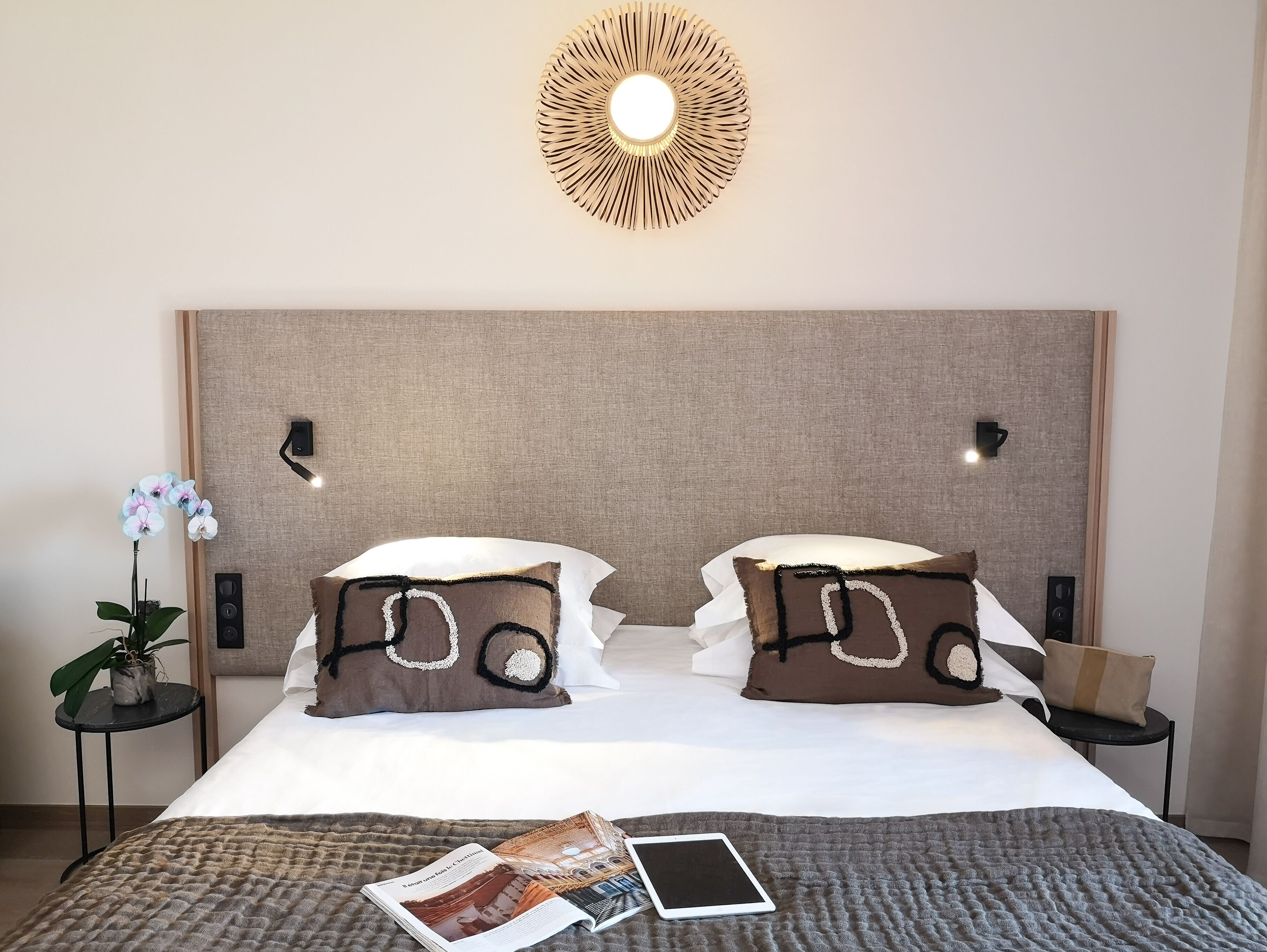 Un lit bien fait avec deux oreillers, une table de chevet avec une lampe, un luminaire fixé au mur et une petite plante en pot.