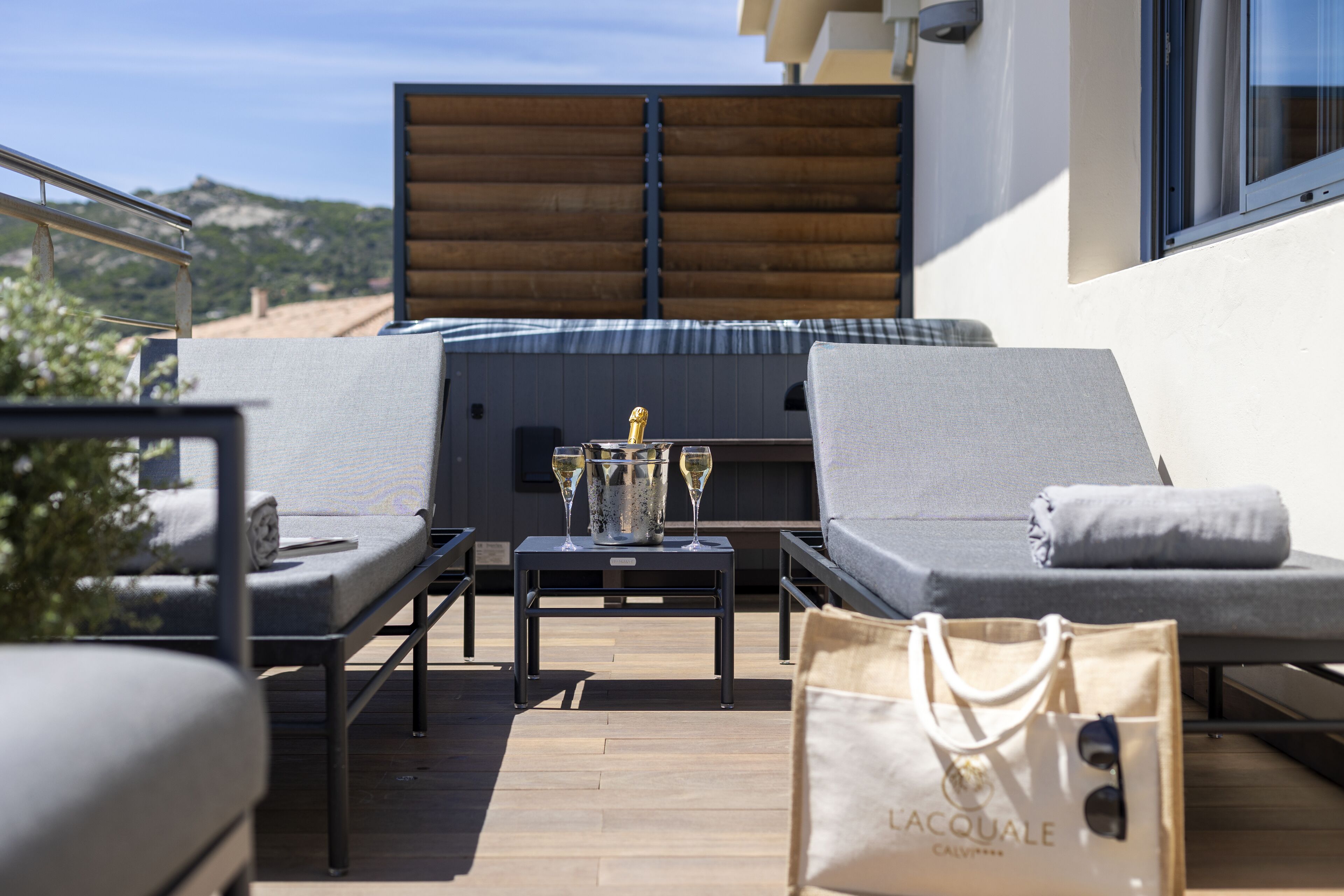 Une terrasse avec deux fauteuils, une petite table avec une bouteille et des verres, et un sac sur lequel on peut voir l’inscription « LACQUALE ».