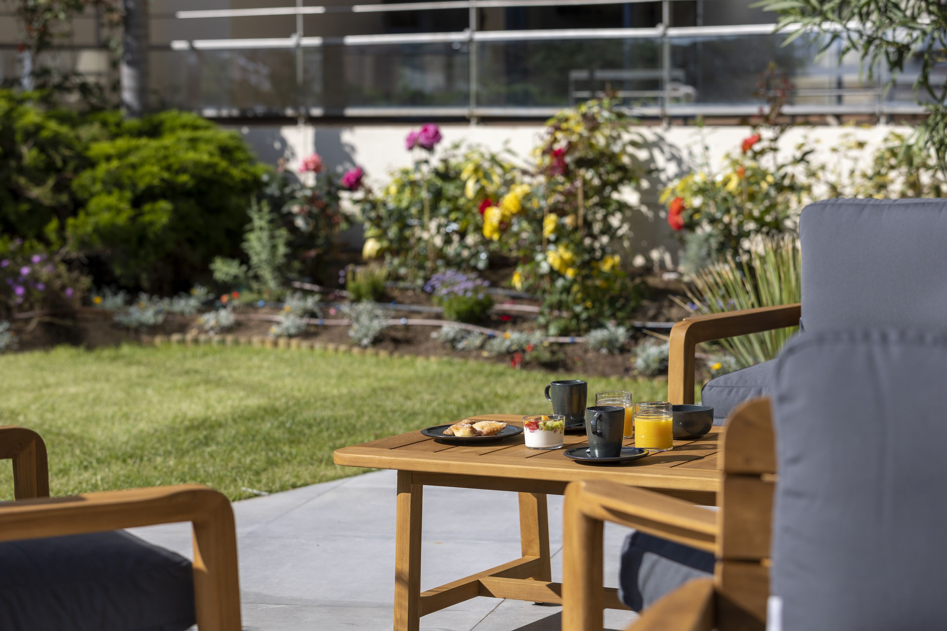 A wooden table with breakfast items and a glass of orange juice, set on a patio with a garden view.