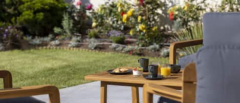A wooden table with breakfast items and a glass of orange juice, set on a patio with a garden view.