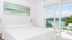 Een hotelkamer met een bed, een schilderij, uitzicht op een meer en bergen, en een balkon met stoelen.