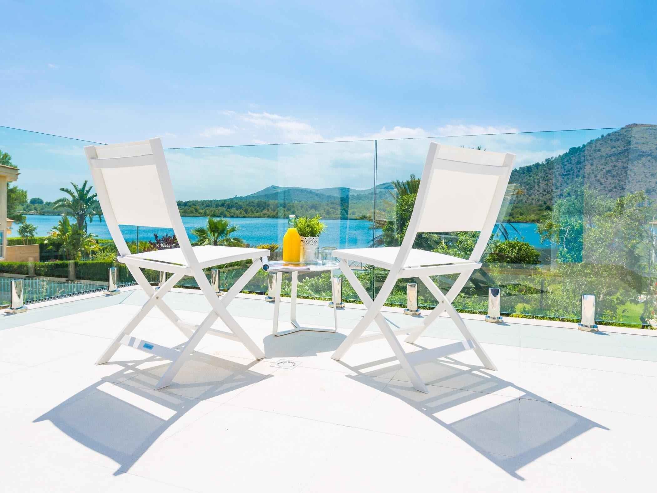 Two white chairs and a small table with a glass of juice on a balcony overlooking a body of water and greenery.