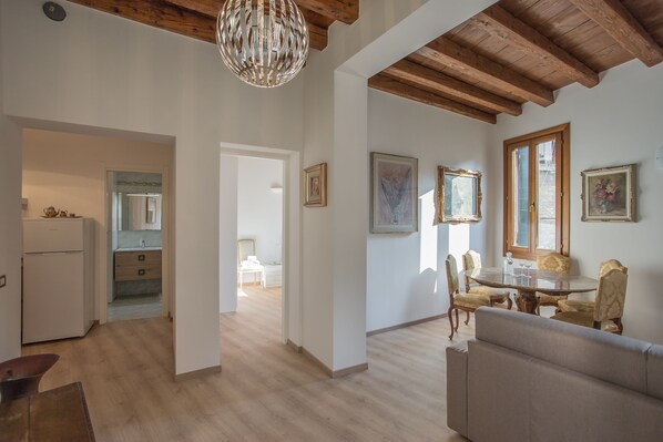 A spacious living area with a dining table, chairs, and a sofa. There is a kitchen visible in the background, and the room has wooden beams on the ceiling.