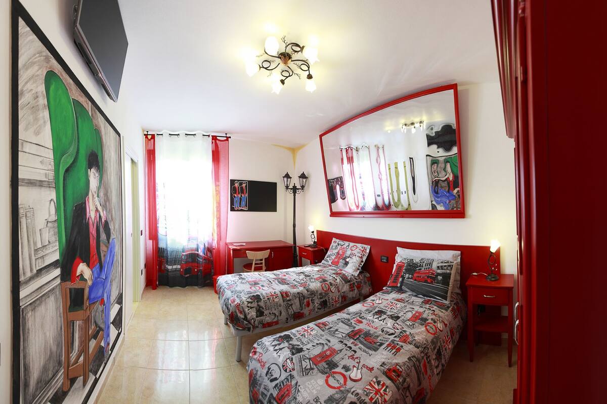 A bedroom with two beds, a desk, a chair, a mirror, and a painting on the wall.