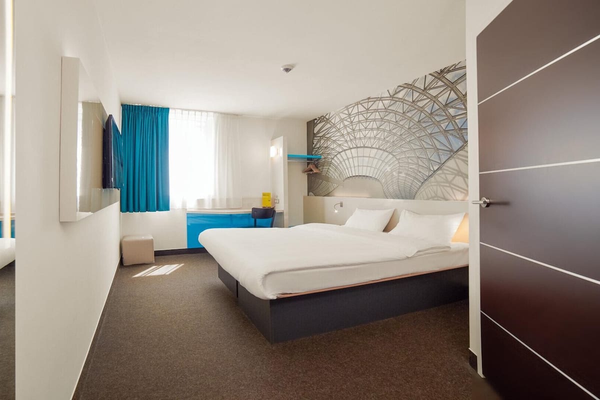 A modern hotel room with a large bed, a desk, a chair, a small table, and a decorative wall piece.