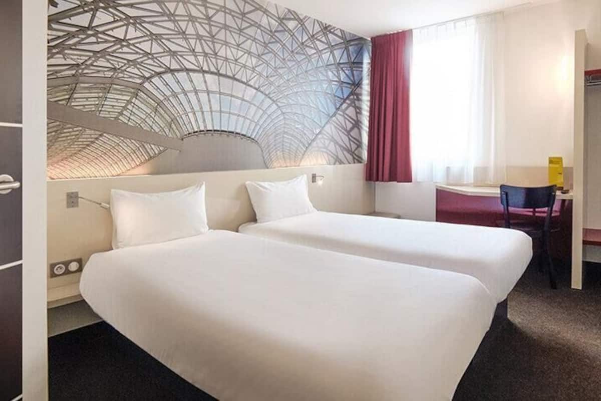 A hotel room with two beds, a desk, a chair, a coat rack, and a wall with a geometric design.