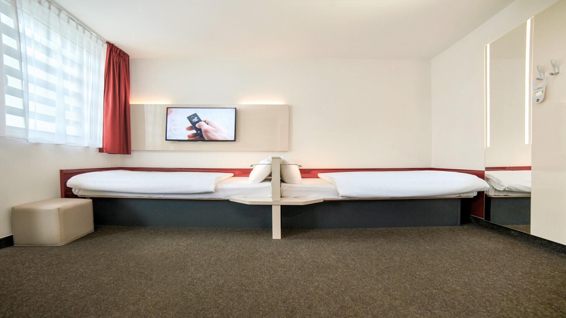 Una habitación de hotel con dos camas, un televisor montado en la pared, una mesita de noche y una cortina roja.