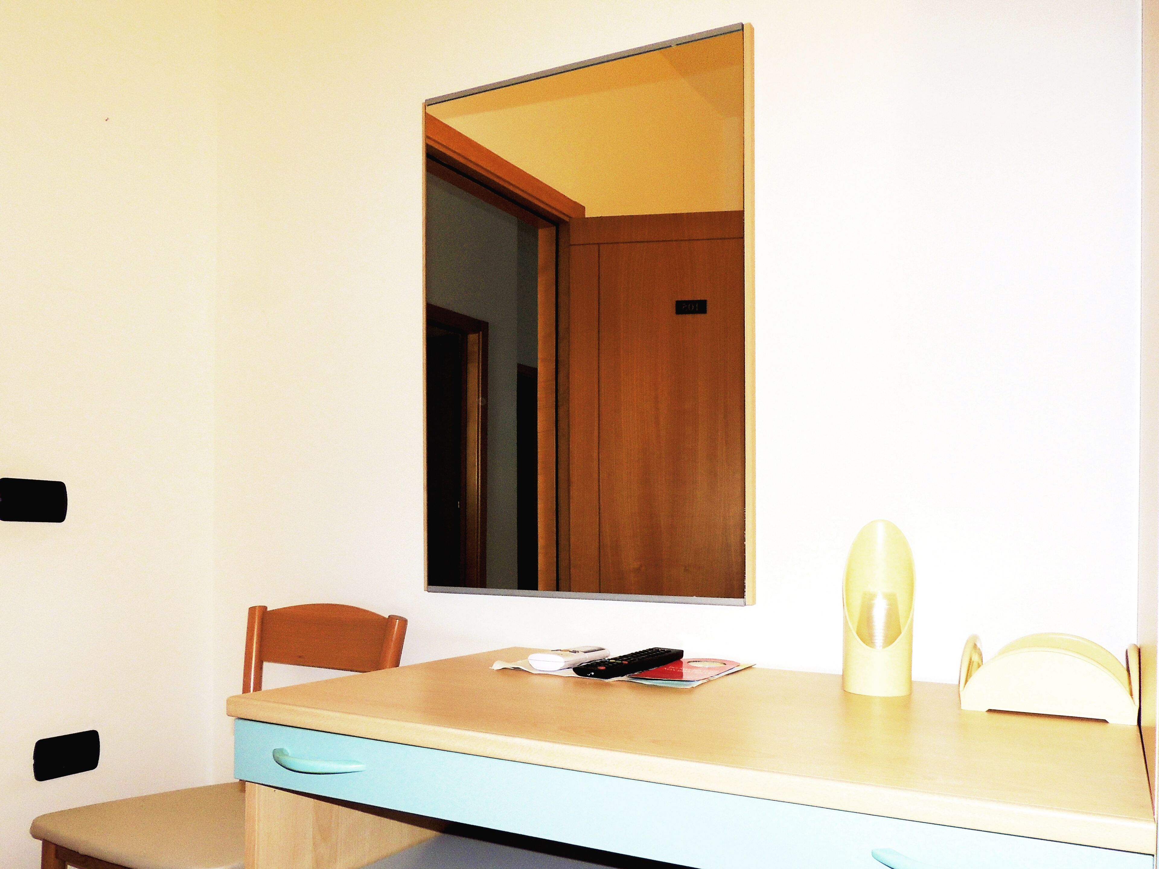 A small room with a wooden door, a single chair, a table with a drawer, and a mirror on the wall.