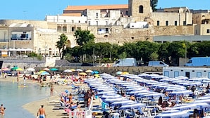 A crowded beach with numerous umbrellas and sun loungers, a stone building with a tower in the background, and a clear blue sky. - Hotel Porto Badisco (Otranto)