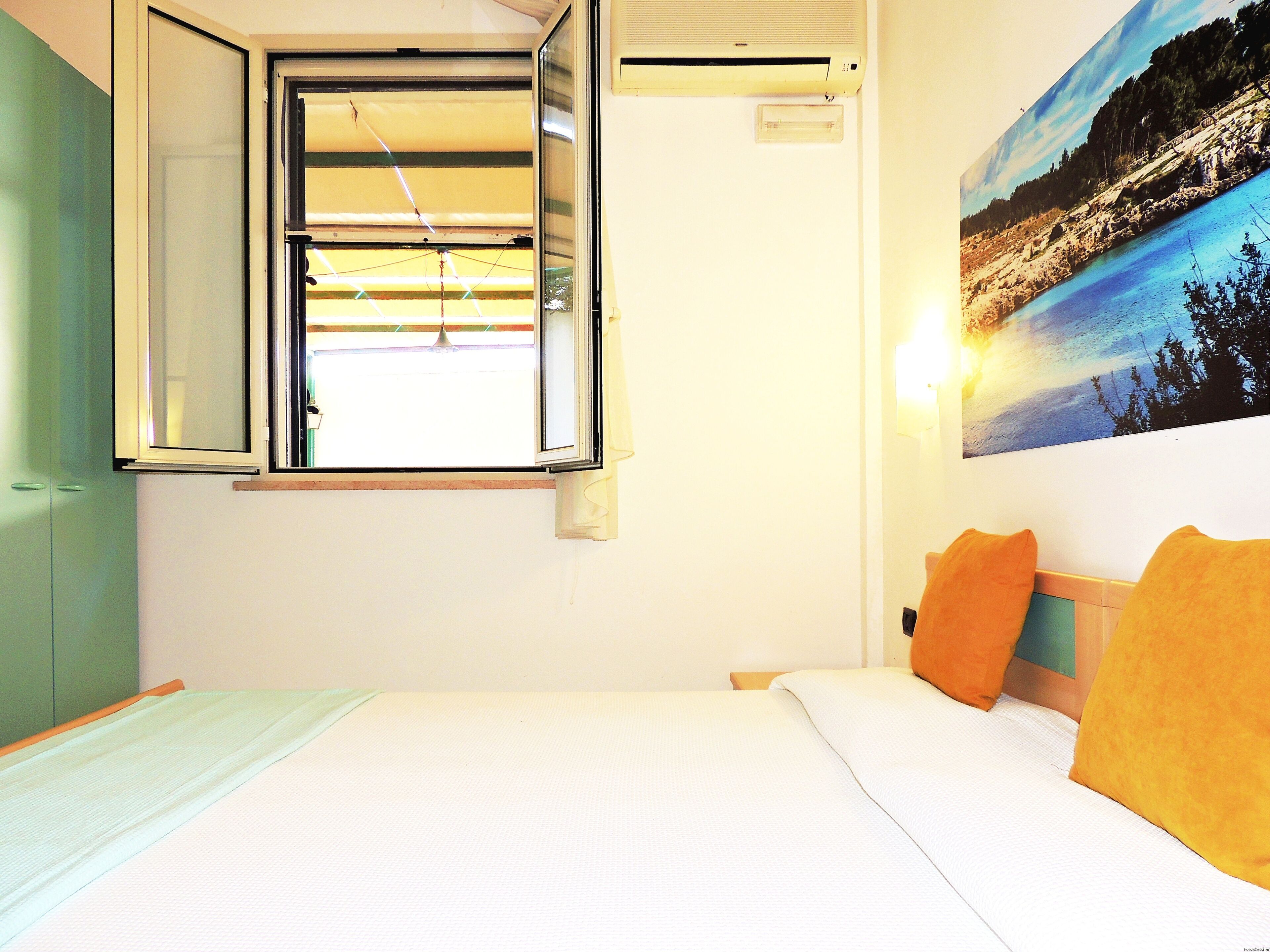 A bedroom with a bed, a window, a wall-mounted air conditioner, and a painting of a landscape.