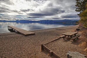Una playa arenosa con un muelle de madera y una mesa de picnic, con vistas a un lago tranquilo y montañas al fondo.