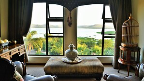 Quarto com uma grande janela com vista para um lago, um baú de madeira, uma gaiola de pássaro e um banco acolchoado.