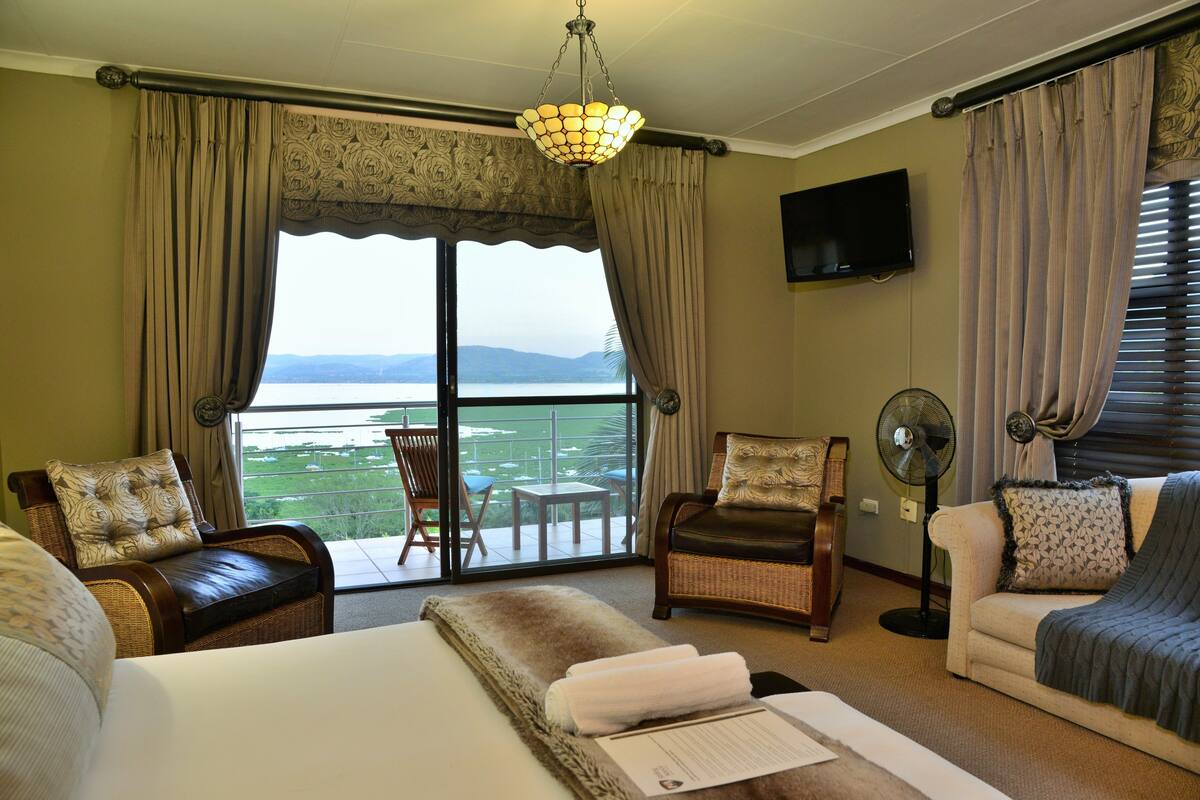 A hotel room with a large bed, two armchairs, a sofa, a balcony with a view, and a television.