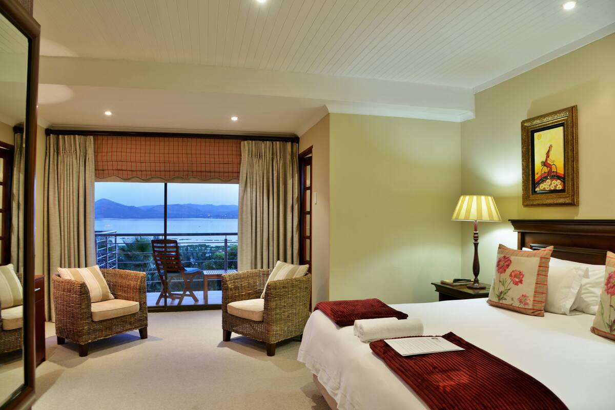 A hotel room with a bed, two wicker chairs, a view of the sea, and a painting on the wall.