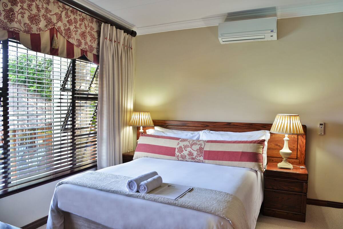 A neatly made bed with a rolled towel and a notepad, a bedside lamp, and a window with blinds and curtains.