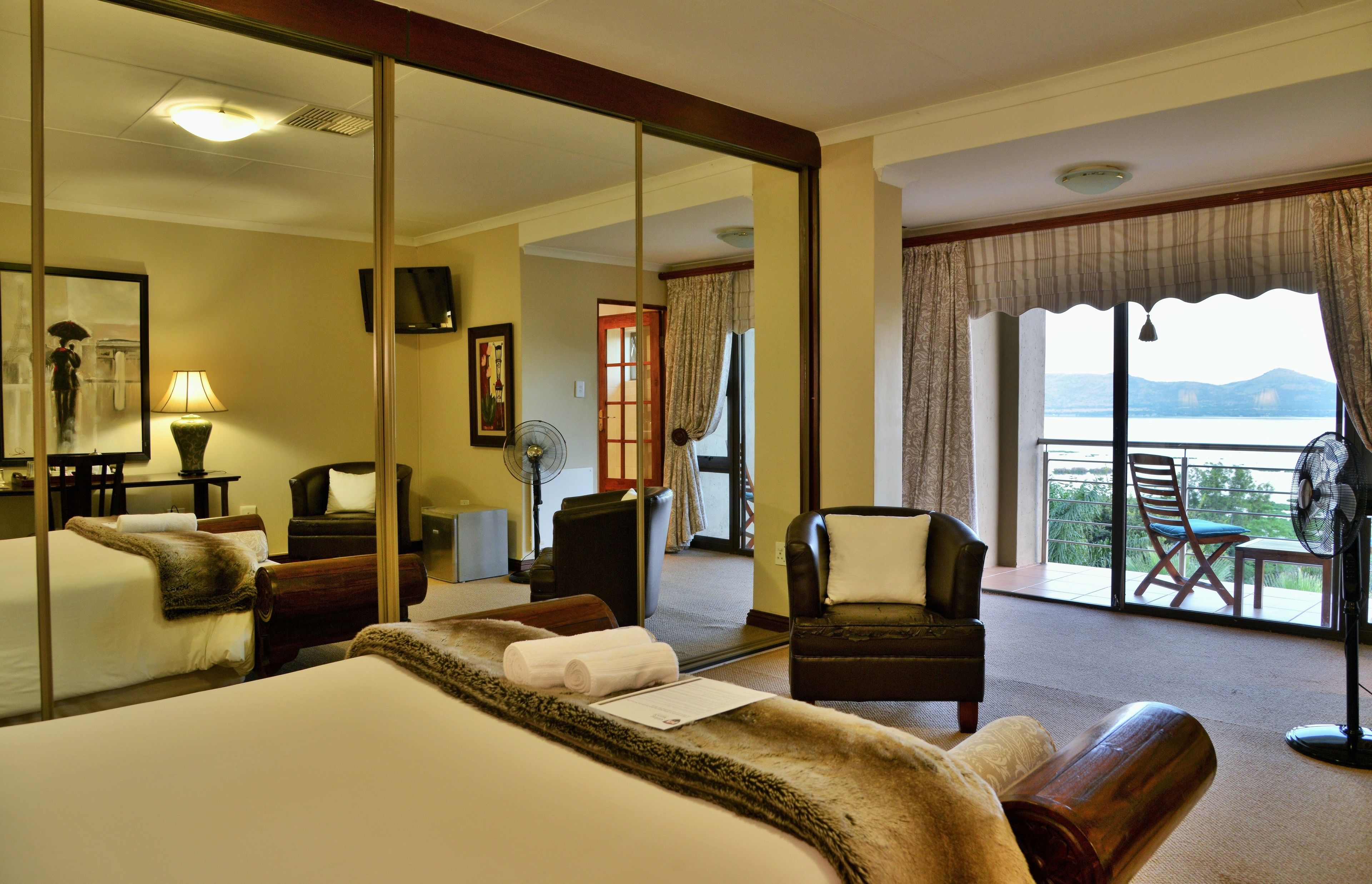 Quarto de hotel com uma cama grande, um espaço de estar com uma poltrona e uma mesa, varanda com vista e um espelho grande refletindo o quarto.