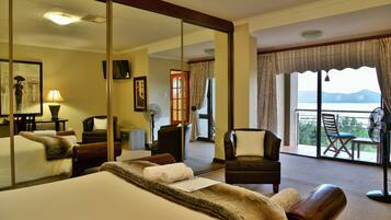 Quarto de hotel com uma cama grande, um espaço de estar com uma poltrona e uma mesa, varanda com vista e um espelho grande refletindo o quarto.