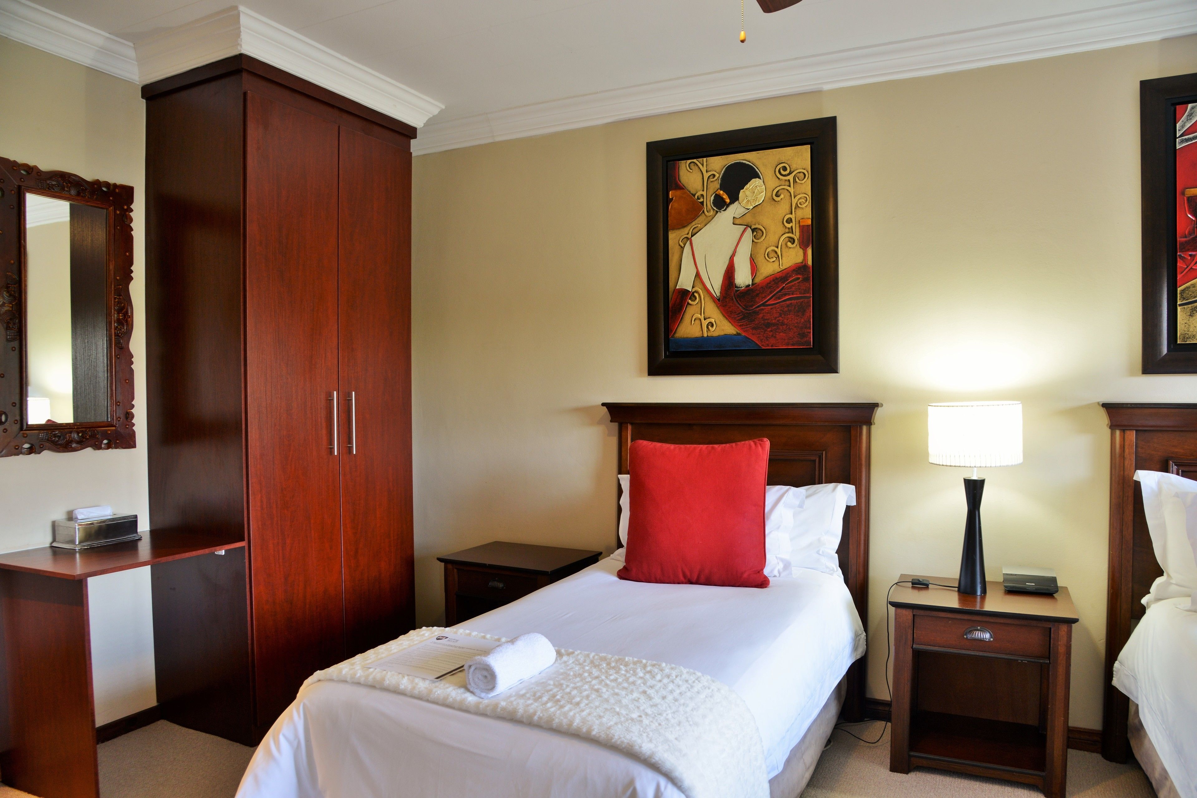 A hotel room with a large wooden wardrobe, a bed with white linens and a red pillow, a nightstand with a lamp, and a framed artwork on the wall.