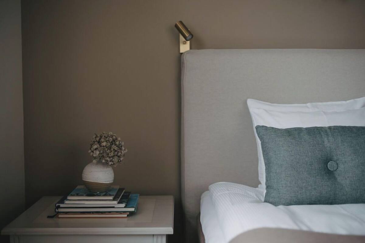 A neatly made bed with white and grey bedding, a small bedside table with a vase of dried flowers and a few books, and a wall-mounted light fixture.
