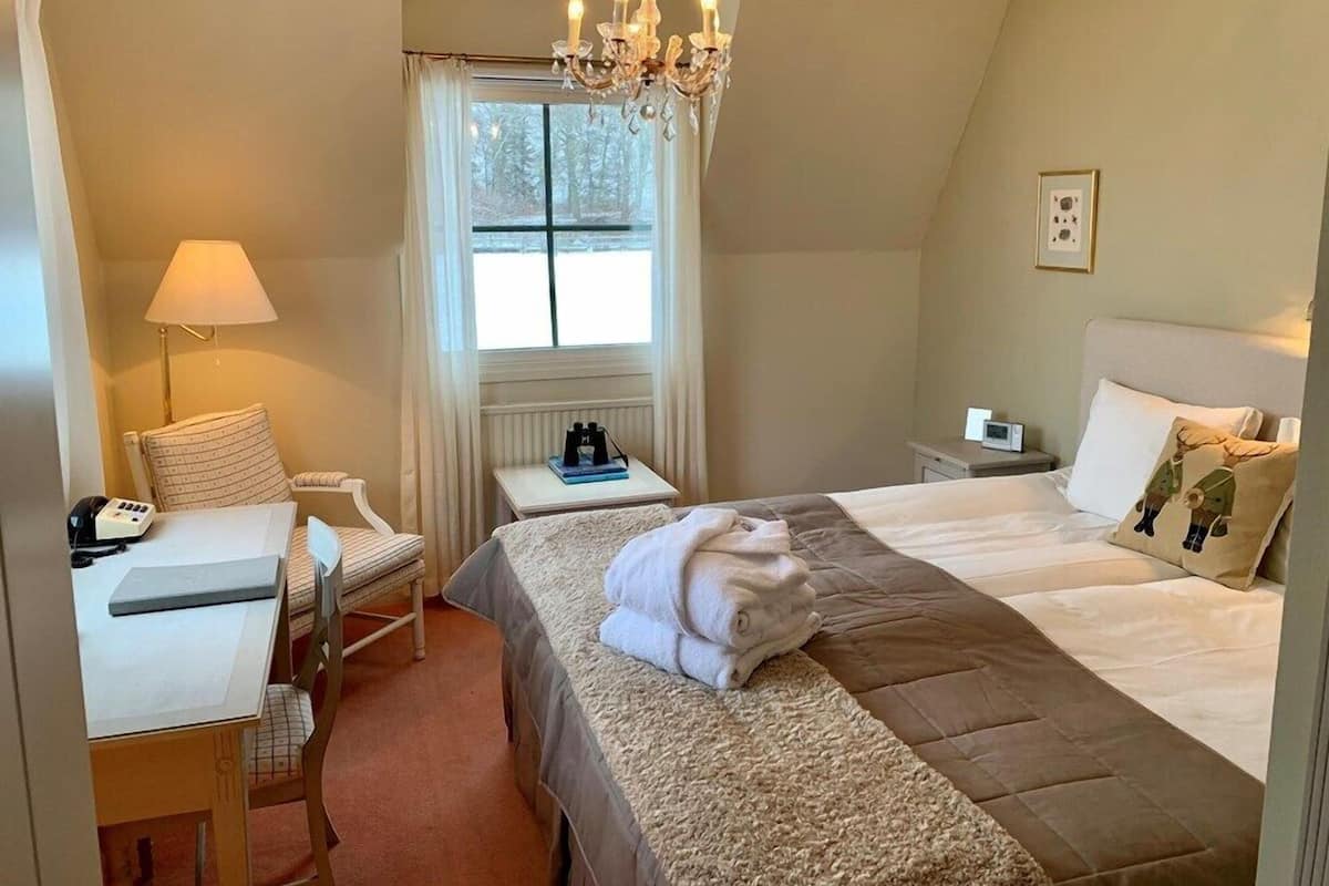 A hotel room with a bed, a desk with a lamp, a chair, a window with curtains, a chandelier, and a bedside table.