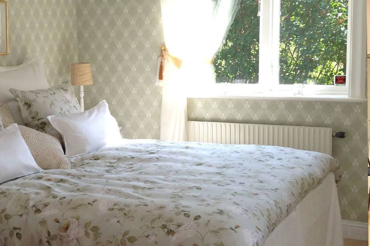 A bedroom with a floral bedspread, multiple pillows, a bedside lamp, and a window with a view of greenery.