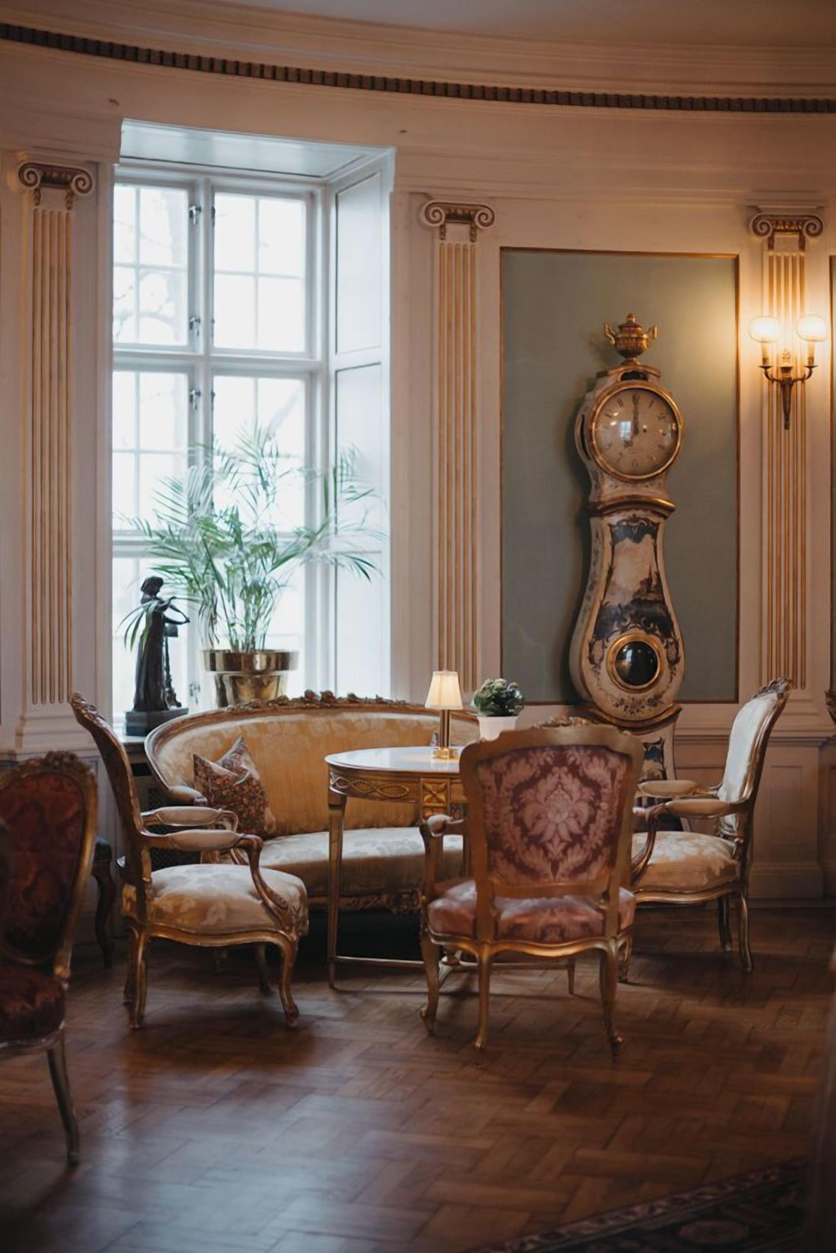 Una habitación con un ventanal grande, un reloj de pie alto y una mesa redonda con sillas.
