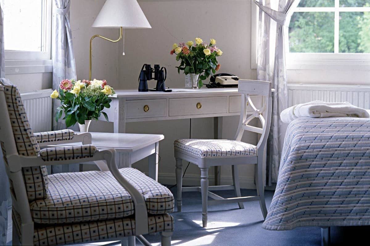 A room with a desk, two chairs, a lamp, a vase with flowers, and a bed.