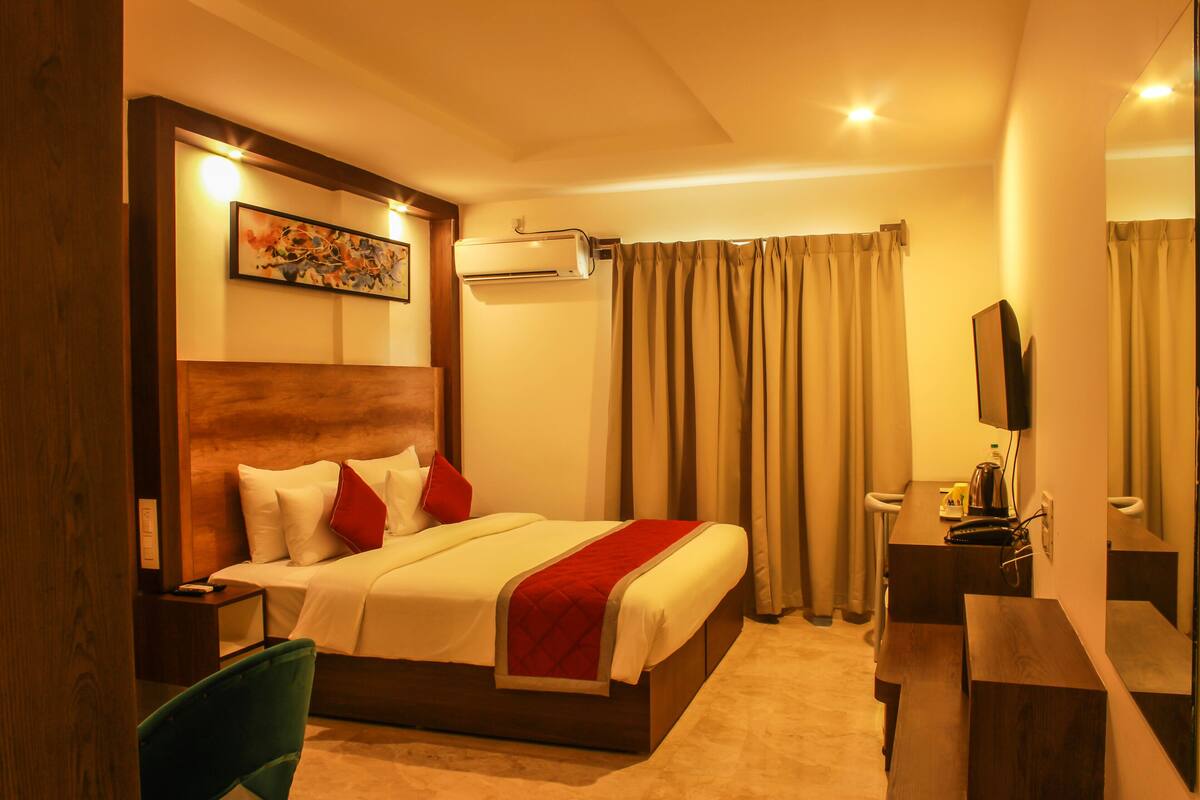 A hotel room with a bed, a desk, a chair, an air conditioning unit, and a television.