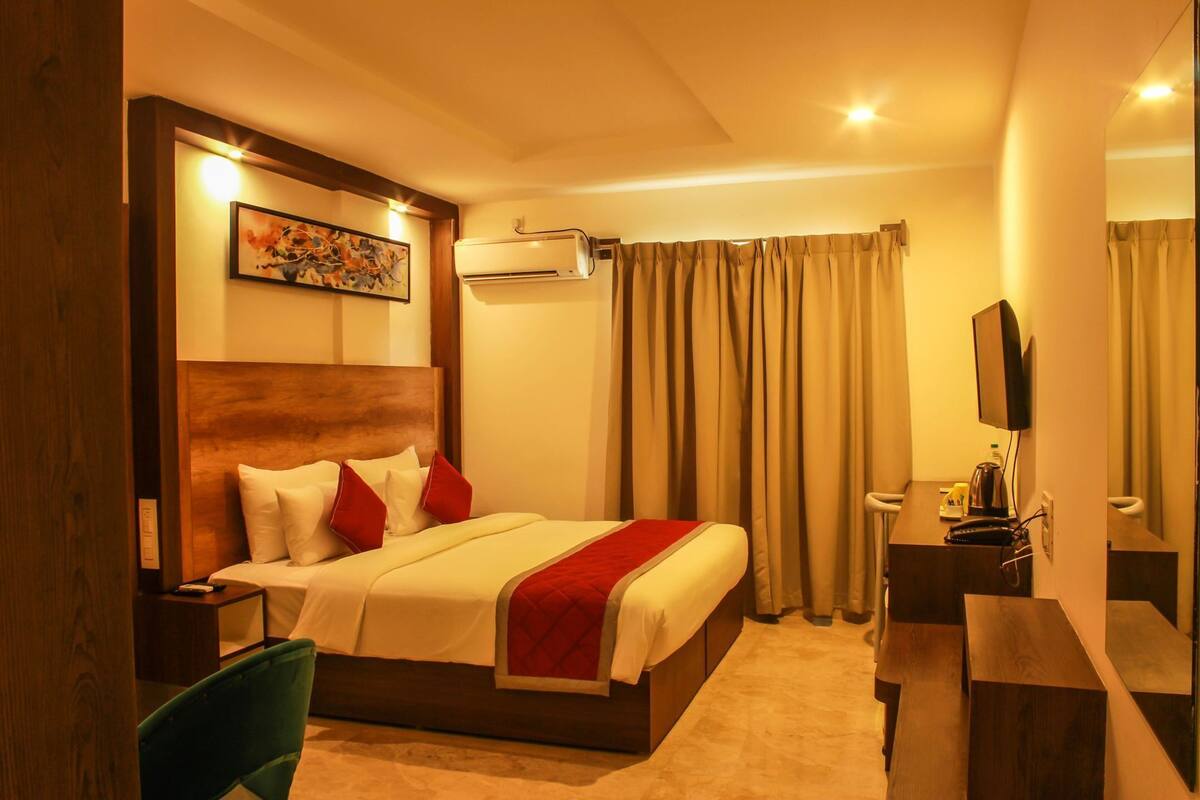 A hotel room with a bed, a desk, a chair, an air conditioning unit, a television, and a painting on the wall.