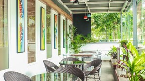 A covered outdoor seating area with wicker chairs and tables, surrounded by large windows and potted plants. - Rest Inn By Sublime (Bengaluru)