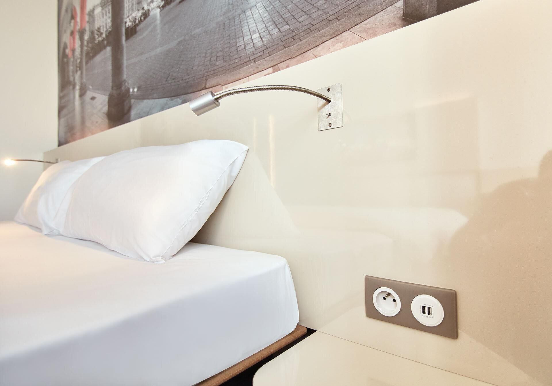 Een hotelkamer met een bed, een wandlamp en een stopcontact met twee aansluitingen.