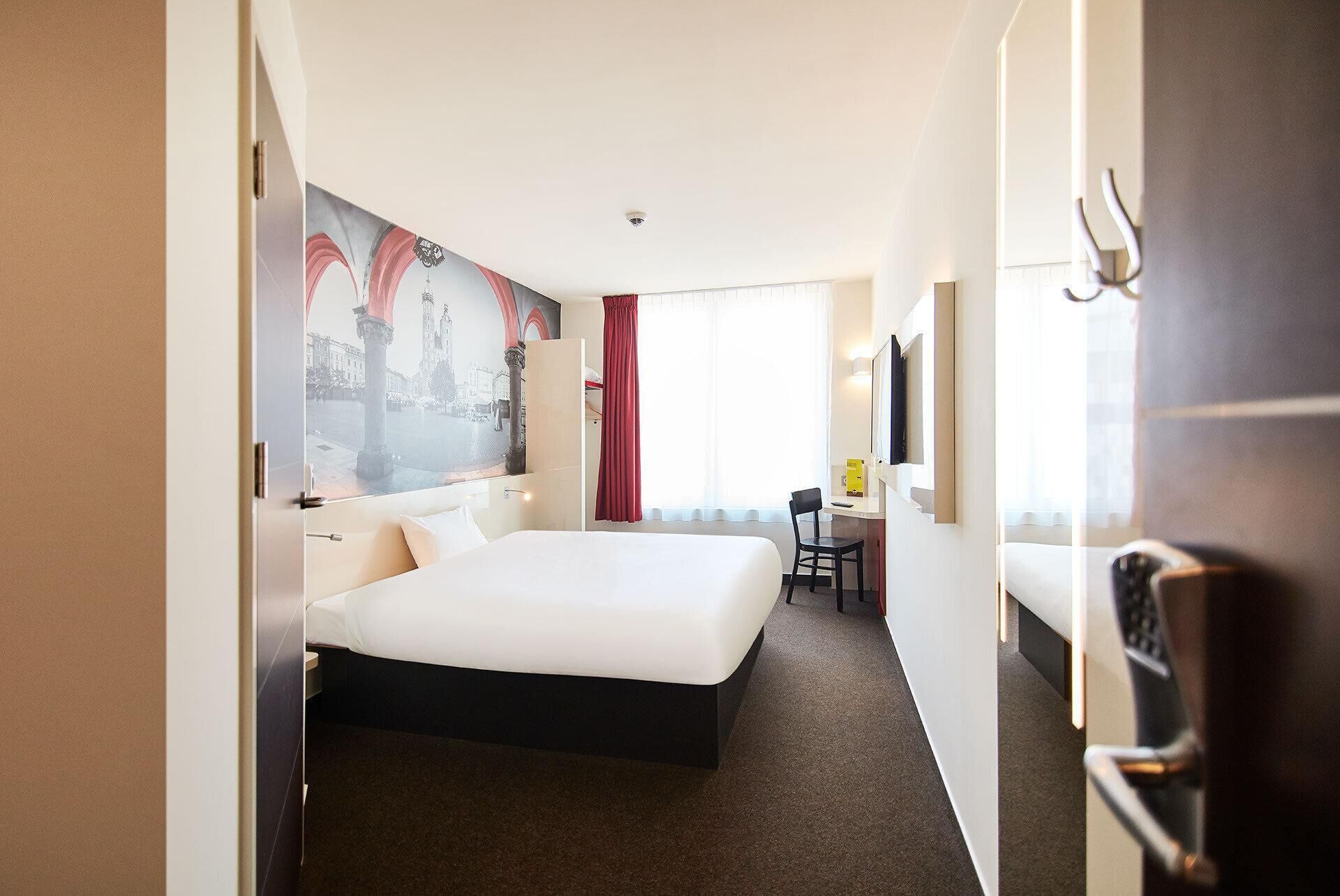 A hotel room with a large bed, a desk, a chair, a TV, and a view of a cityscape through a window.