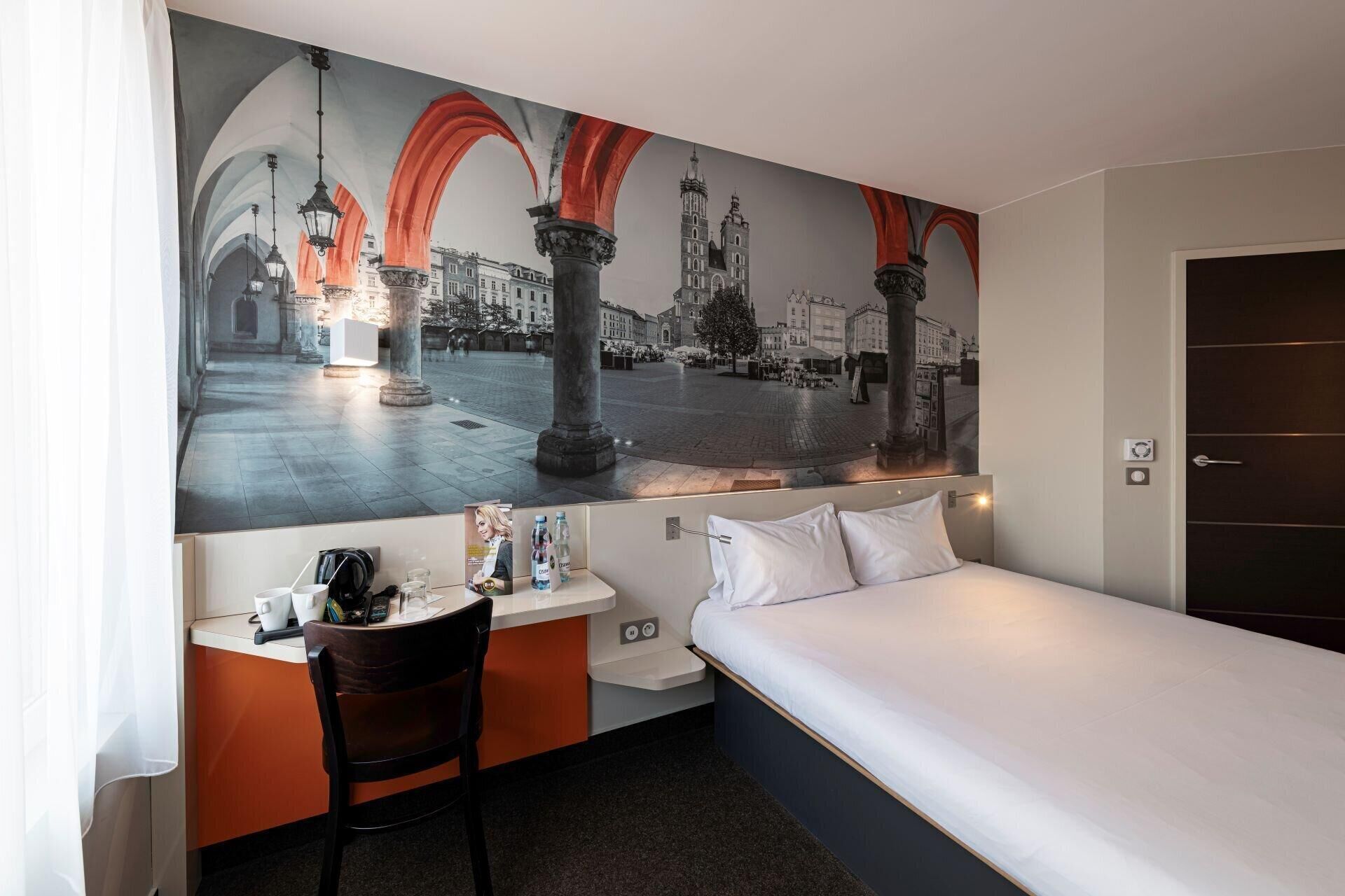 Une chambre d’hôtel avec un lit, un bureau, une chaise et une grande fresque murale représentant un paysage urbain.