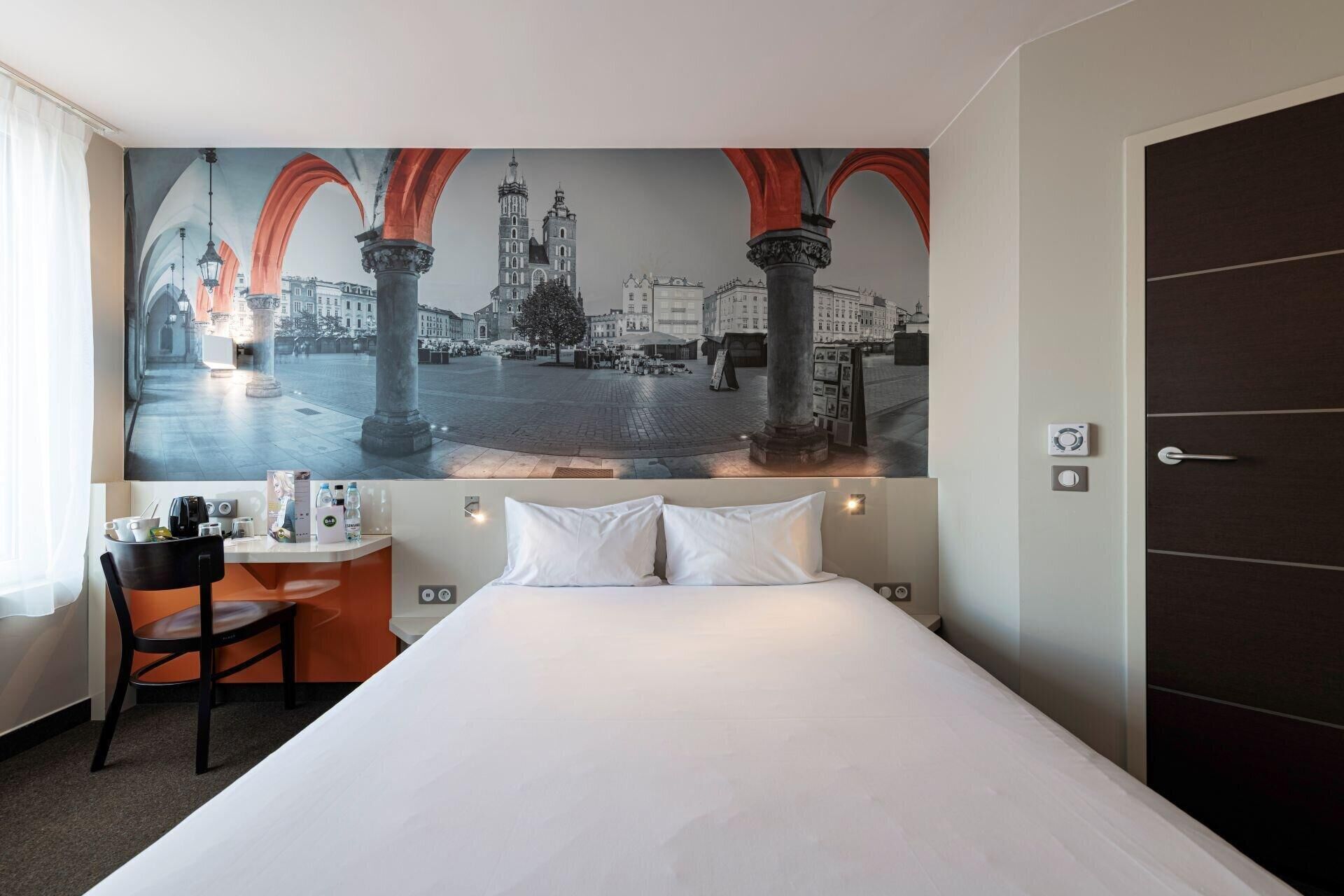 Habitación de hotel con una cama grande, un escritorio con silla y un cuadro en la pared que muestra un paisaje urbano con arcos.