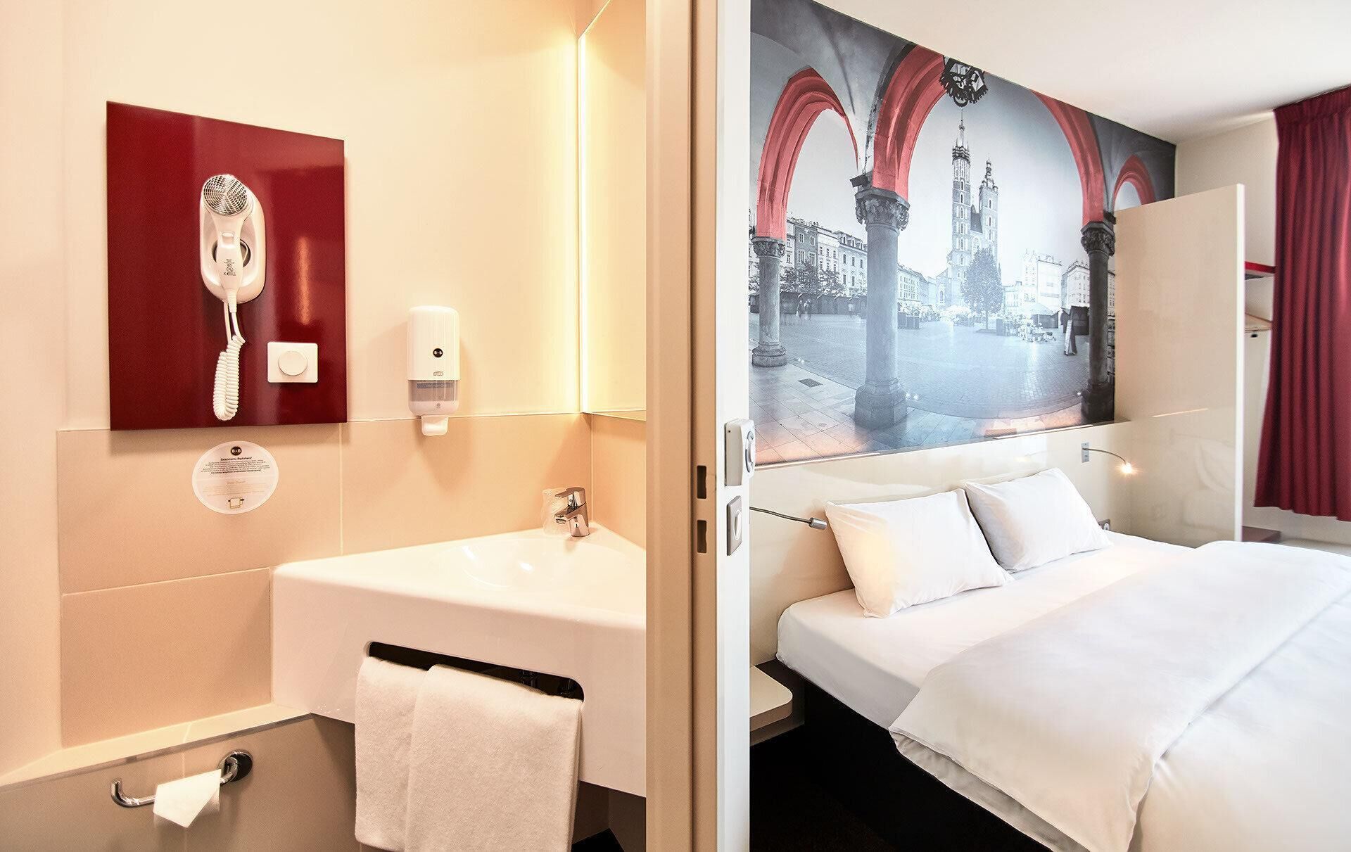 A hotel room with a bathroom featuring a sink, mirror, and towel rack. The room has a bed with white bedding and a view of a cityscape through a window.