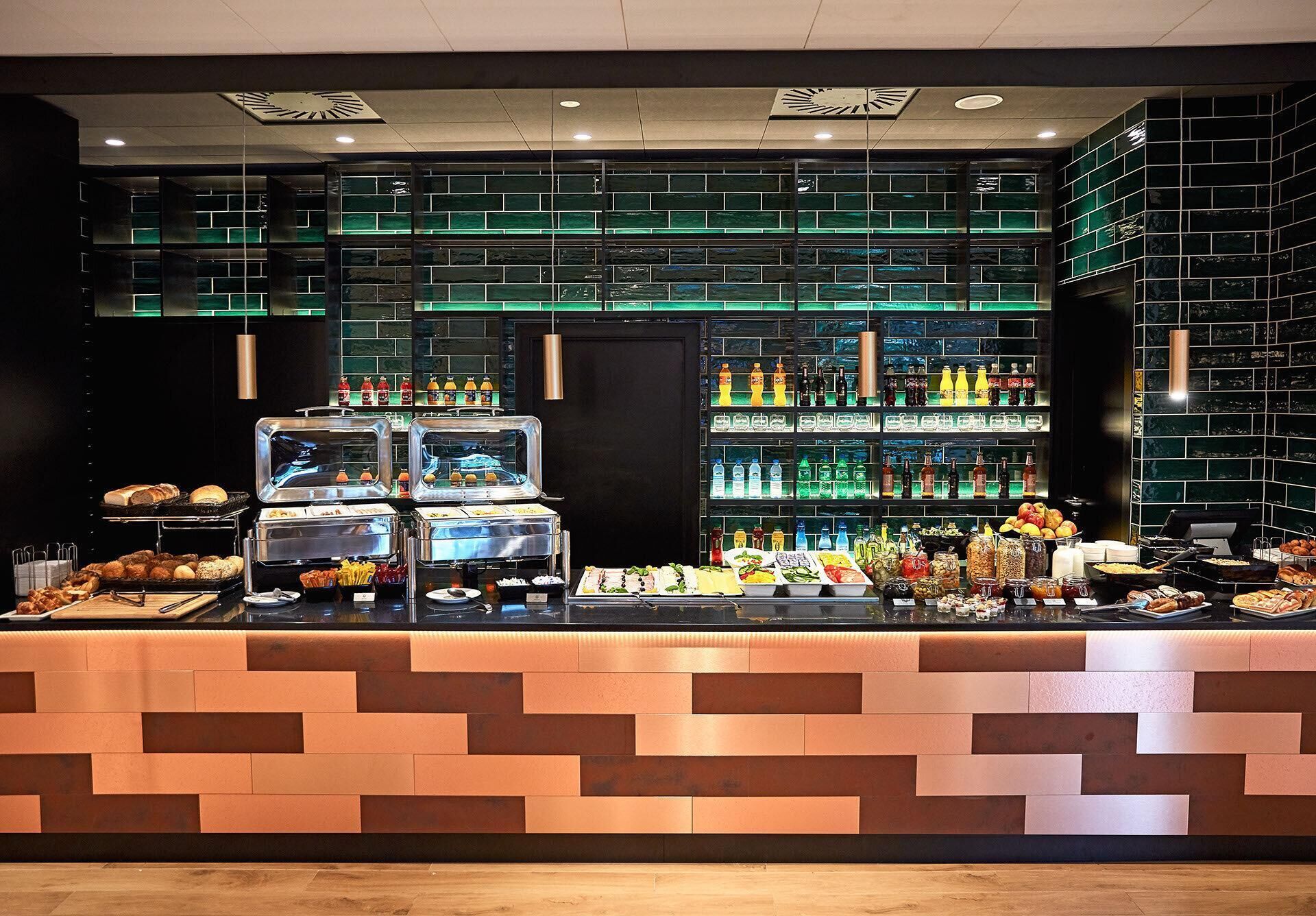 Zona de bufé con diversas opciones de comida y bebidas, un mostrador de diseño moderno, y una pared verde con azulejos que alberga estantes llenos de botellas.