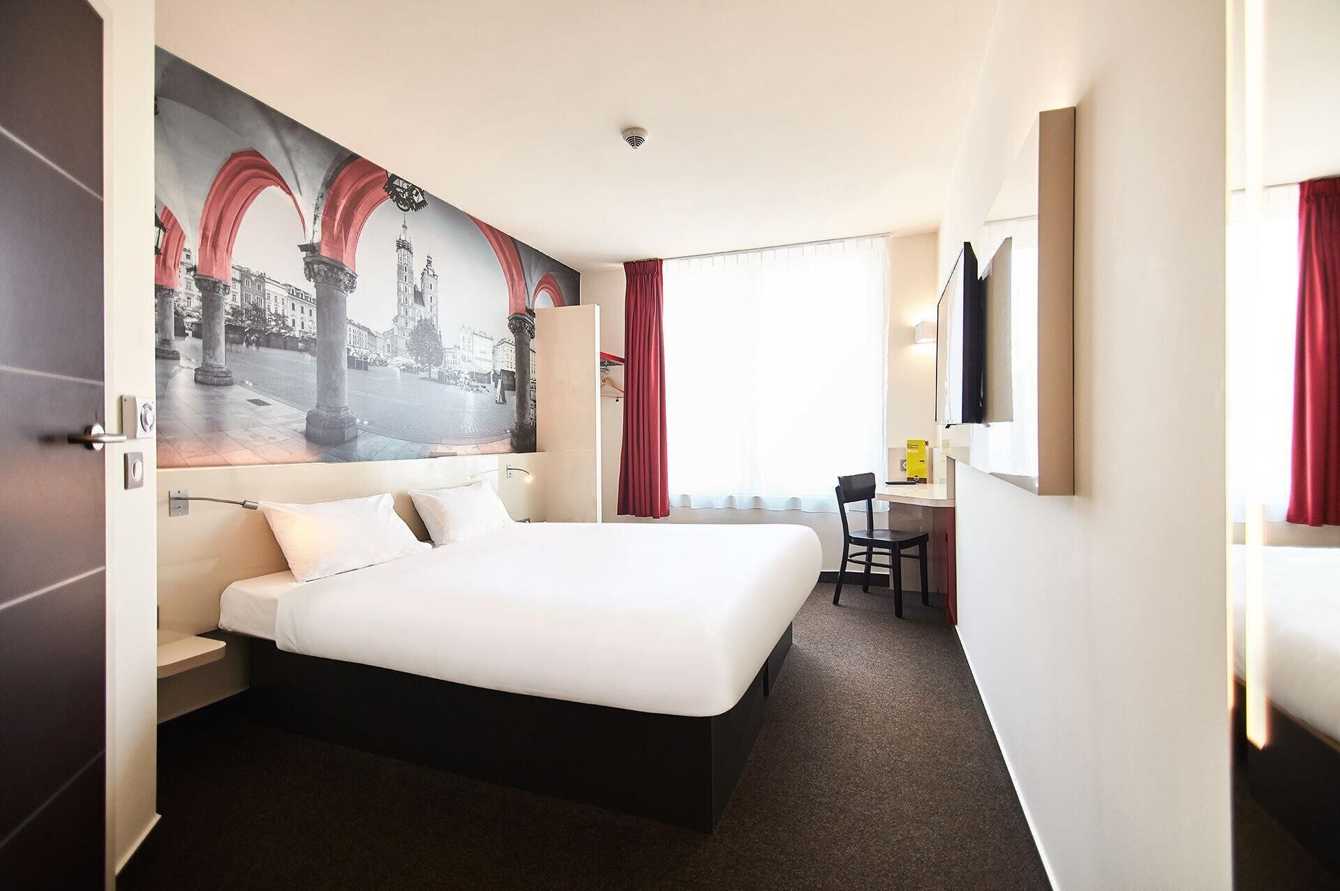 A hotel room with a large bed, a desk, a chair, a TV, and a mural of a cityscape.