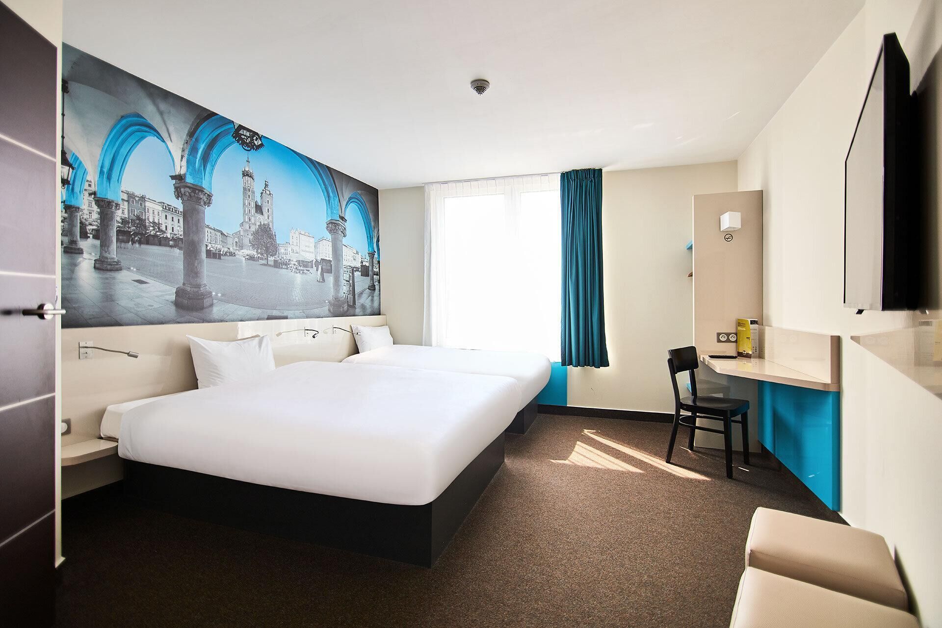 Habitación de hotel con una cama grande, un escritorio, una silla, un televisor y una ventana con cortinas.