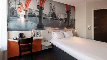A hotel room with a bed, a desk with a chair, a large cityscape mural on the wall, and a door.