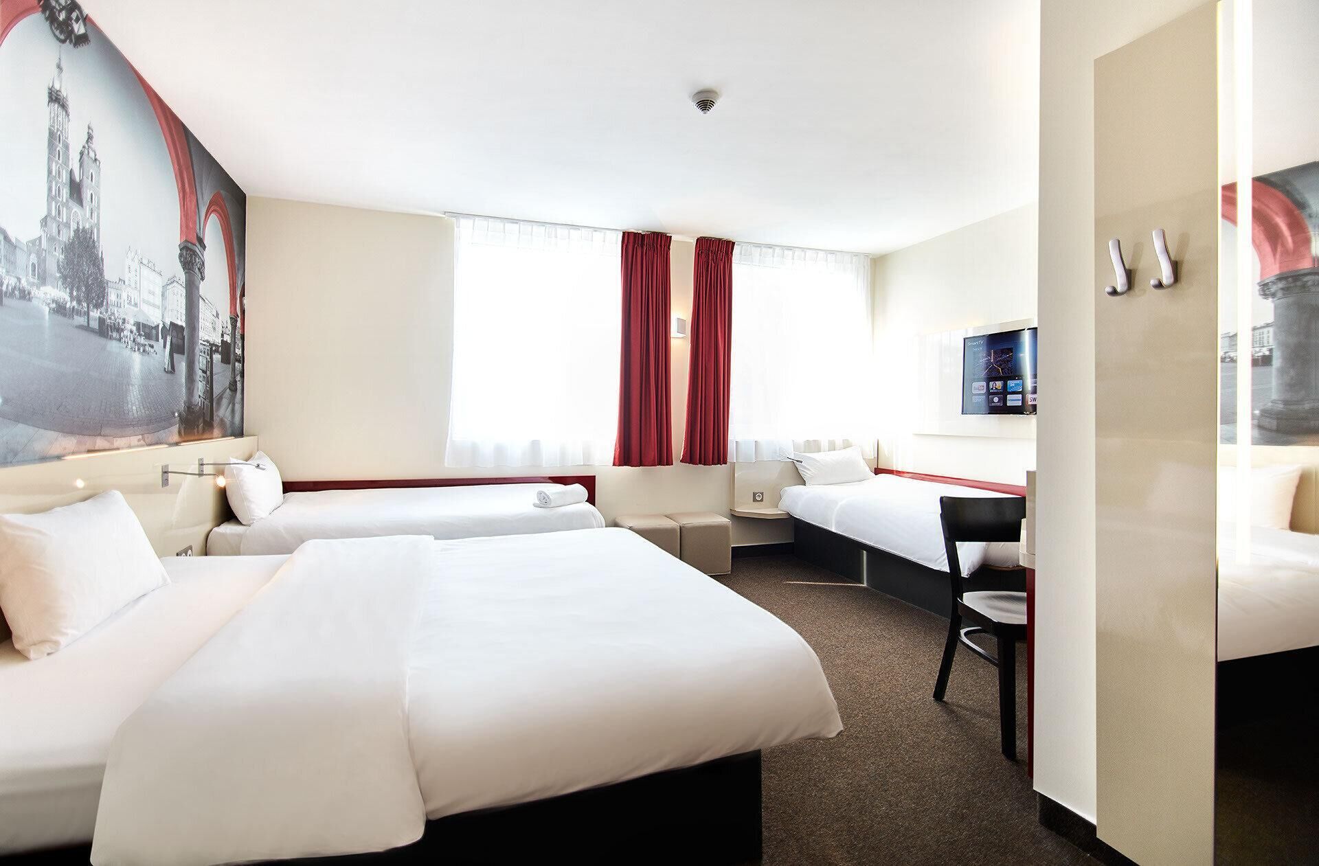 A hotel room with two beds, a desk, a chair, and a large window with red curtains.