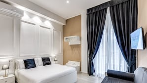 A modern hotel room with a large bed, a bedside table with a lamp, a TV mounted on the wall, and a window with dark curtains. - RAGGIO DI SOLE - ATAROOMS Collection (Naples)