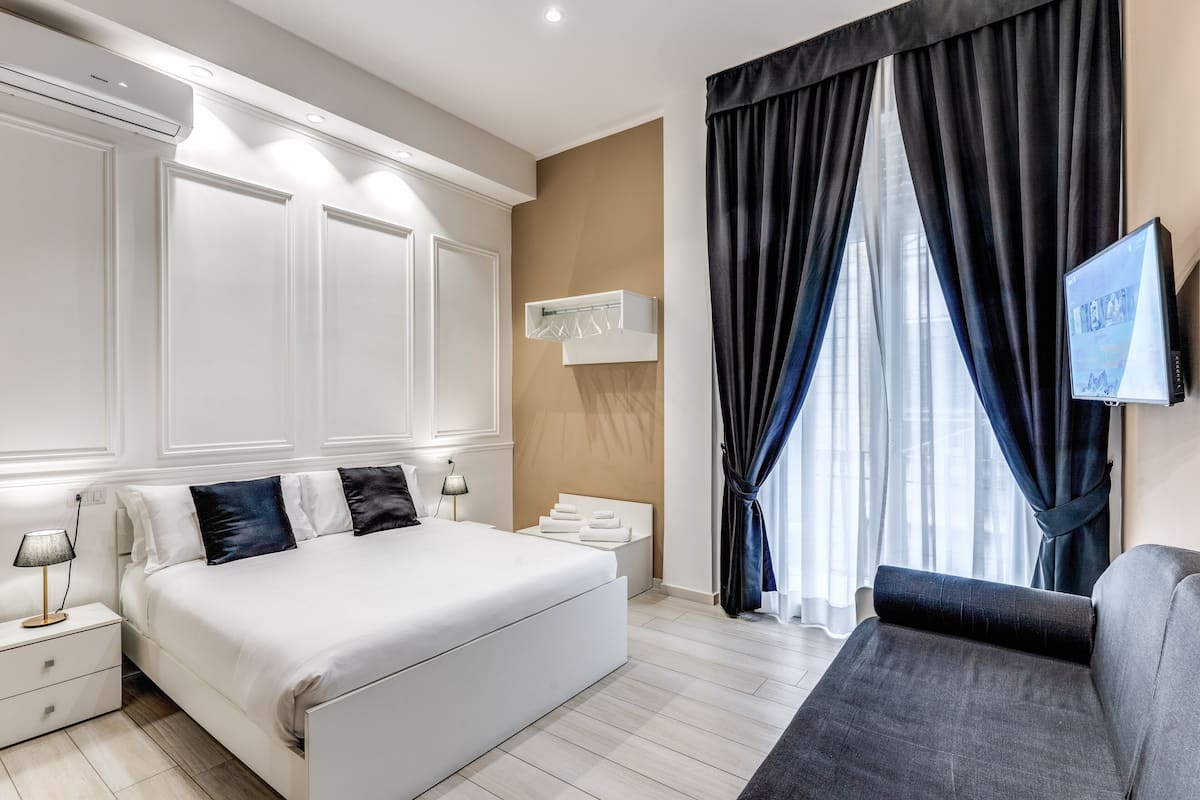 A modern hotel room with a large bed, a bedside table with a lamp, a TV mounted on the wall, and a window with dark curtains.