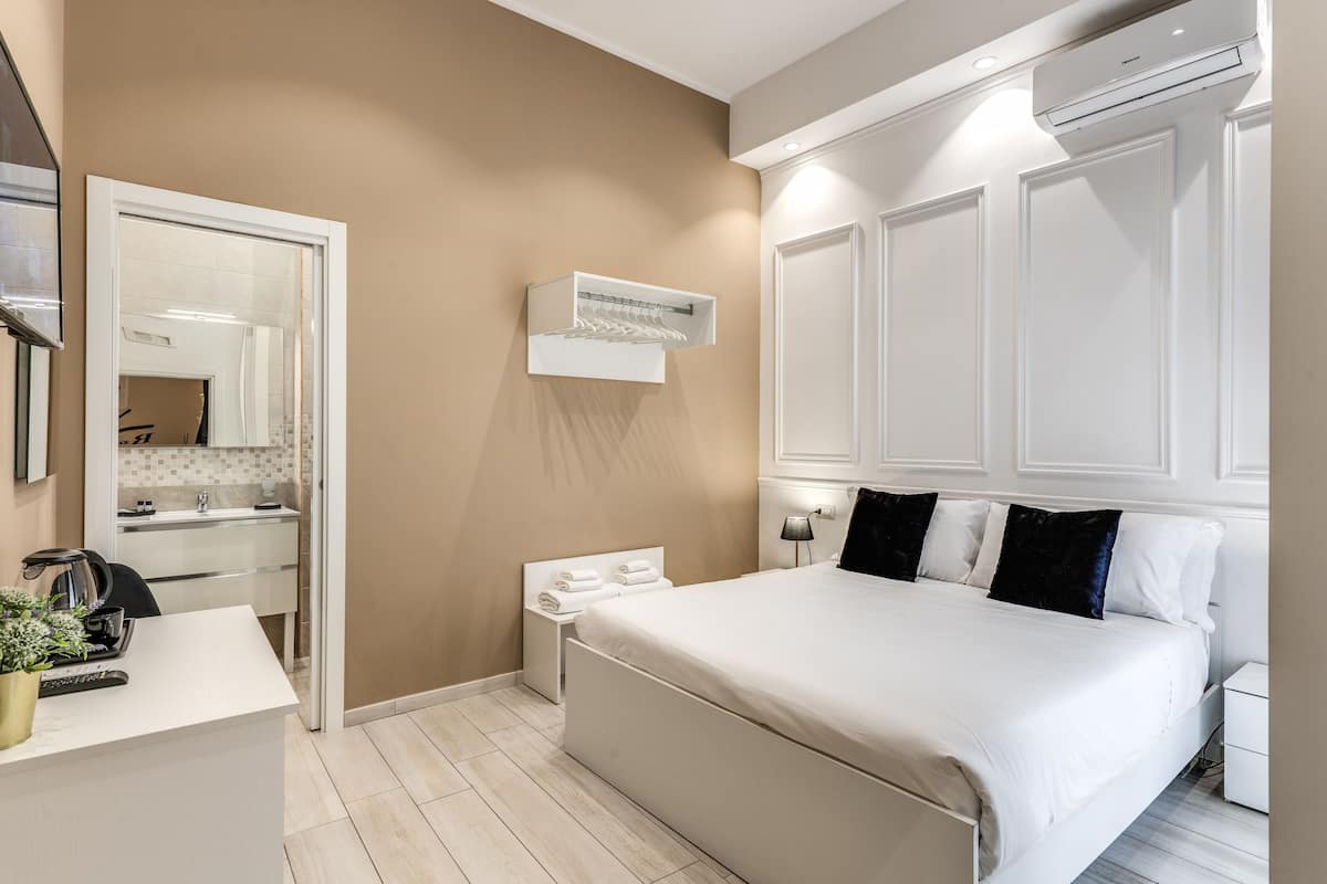 A modern hotel room with a large bed, a bedside table, a mirror, a wall-mounted heater, and a small seating area with a chair and a plant.